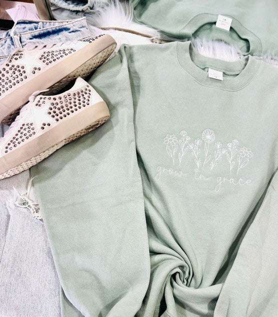 A green embroidered sweatshirt with 'wildflowers' text and a floral pattern, displayed flat with a pair of white sneakers and a denim jacket in the background.