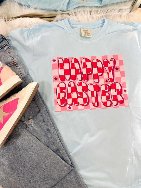 A light blue colored t-shirt with a 'Happy Easter' text and pink checkered pattern on it, displayed on a flat surface with denim jeans and a wooden object in the background.