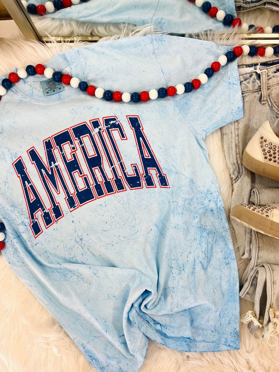 A blue colored graphic tee with the text 'AMERICA' printed in red and white, displayed on a faux fur surface with jeans and a decorative lei in the background.