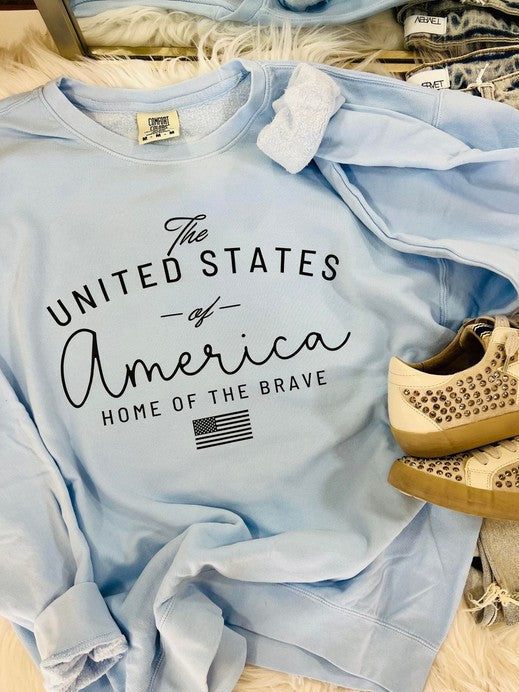 A light blue t-shirt with the text 'The United States of America Home of the Brave' printed on it, displayed flat with a pair of beige shoes at the bottom of the frame.