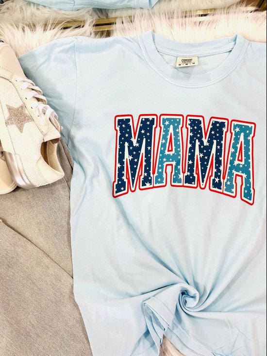A blue curved-neck t-shirt with the word 'MAMA' and a star graphic in red and blue colors, displayed flat with a pair of white sneakers and a furry white blanket in the background.