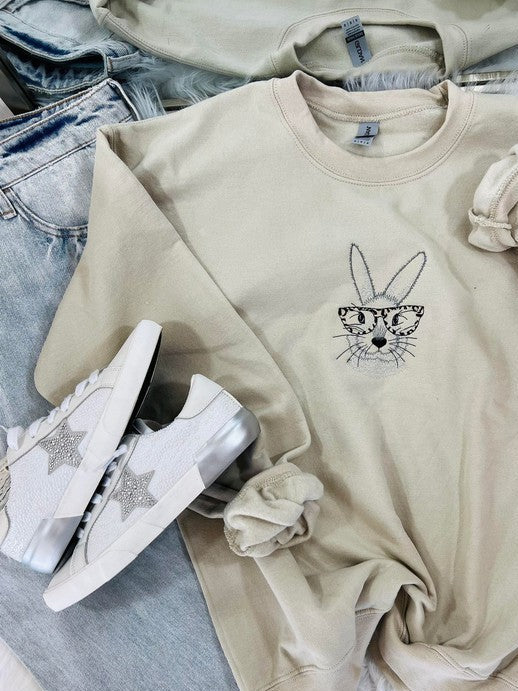 A sand-colored embroidered sweatshirt with a graphic of a bunny wearing glasses, paired with white sneakers and blue jeans.