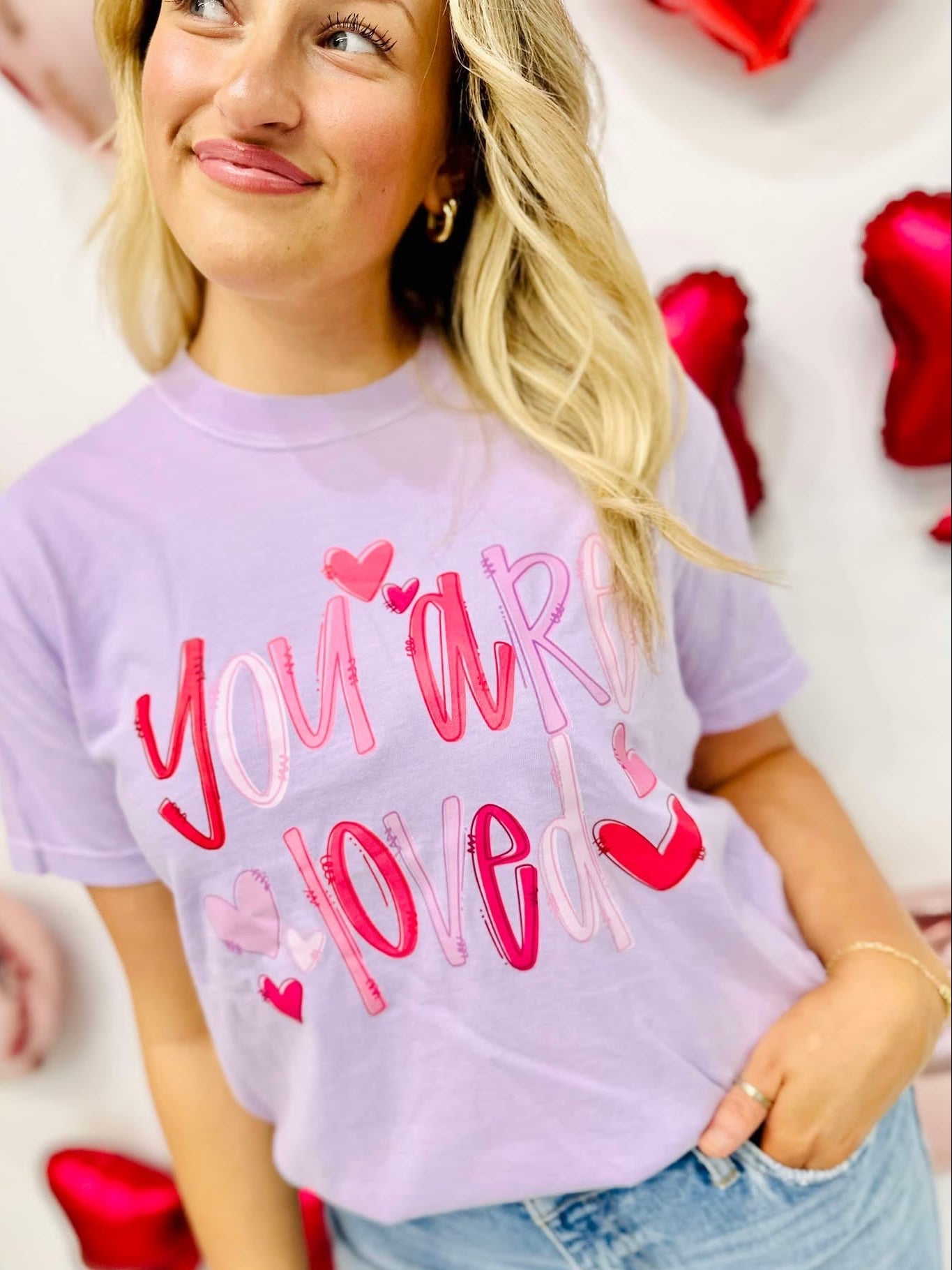 Woman wearing a purple t-shirt with 'You are love' text and hearts, surrounded by red heart-shaped props.
