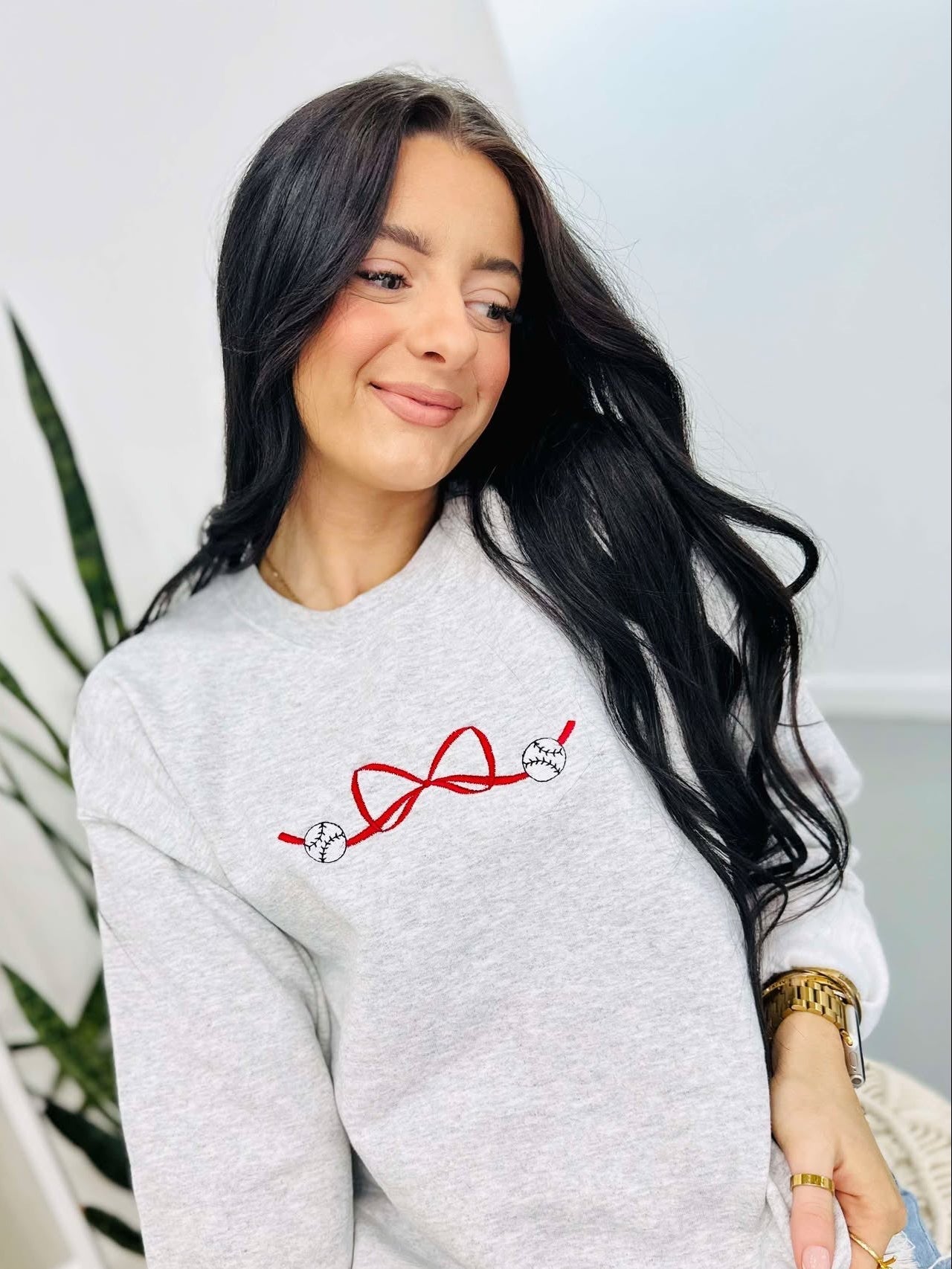 A woman smiling and sitting down, wearing a grey sweatshirt with a red and white embroidered bow design and the baseballs.