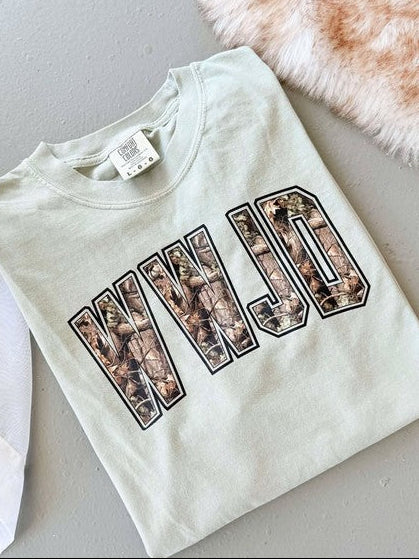 A white t-shirt with 'WWJD' camouflage pattern printed on it, displayed flat with a tag and a furry item in the background.