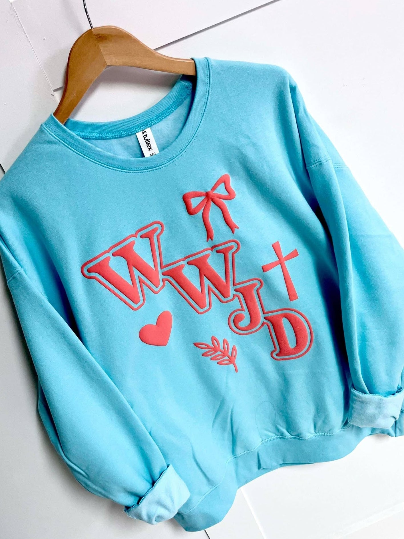 Light blue sweatshirt with coral puff text WWJD and cross, bow, heart leaf graphics on a white tiled floor