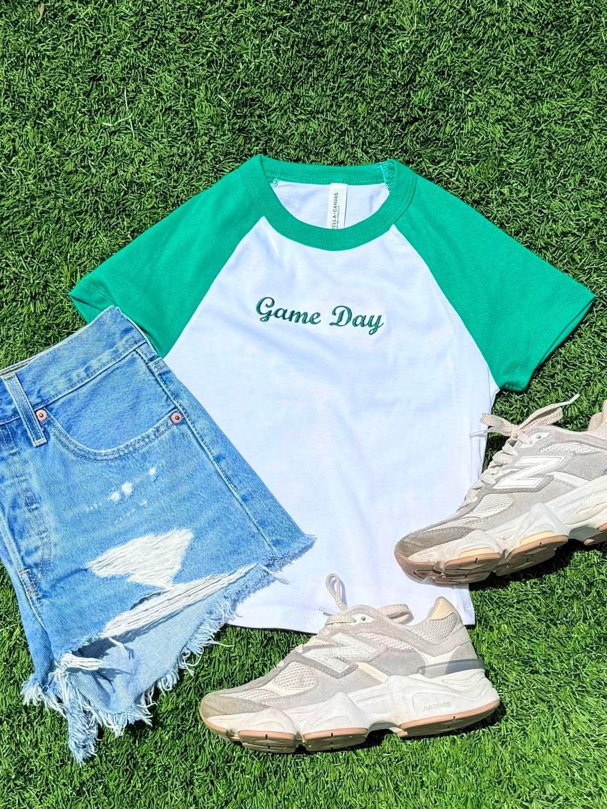 A styled flat-lay of a green and white raglan t-shirt with 'Game Day' printed on the chest, paired with distressed denim shorts, beige sneakers, and arranged on a green turf background.