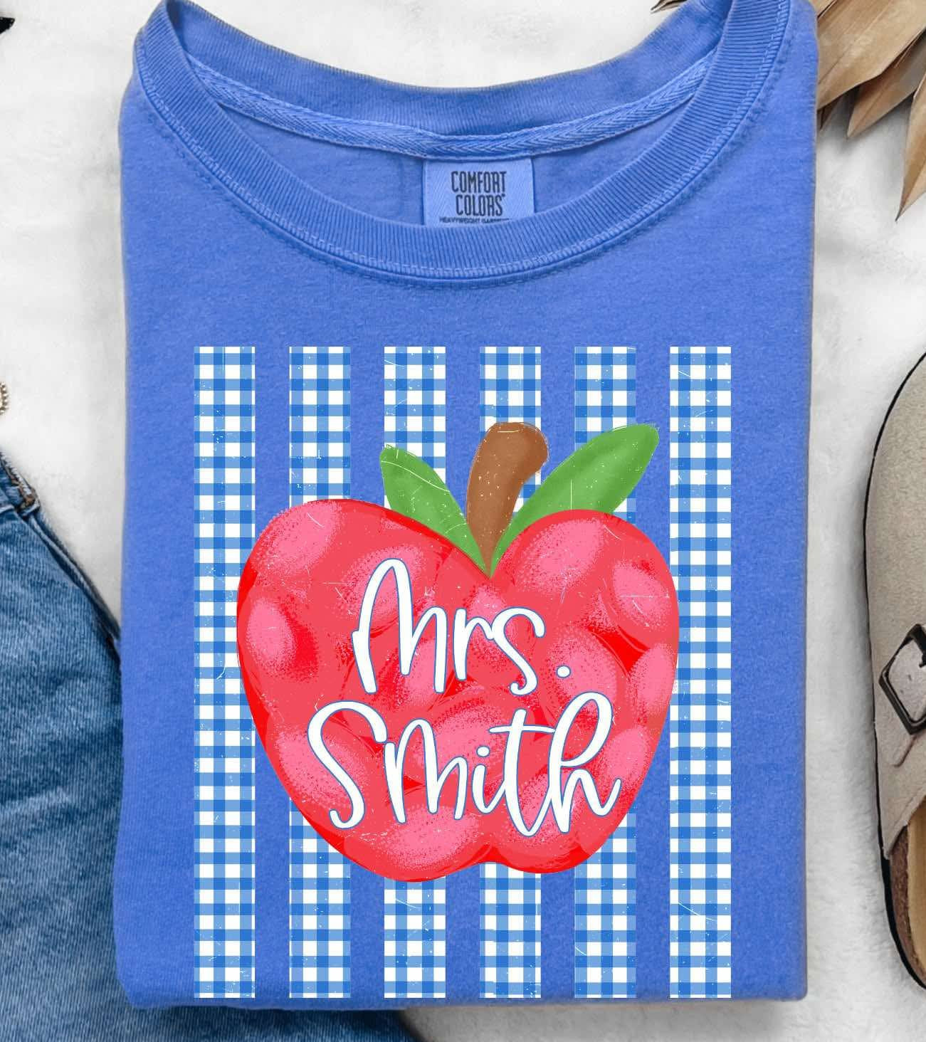A folded blue T-shirt with a red apple graphic and the text 'Mrs. Smith' over blue-and-white gingham stripes, styled in a flat lay with jeans, sunglasses, a necklace, a slip-on shoe, and dried palm leaves on a white textured background.
