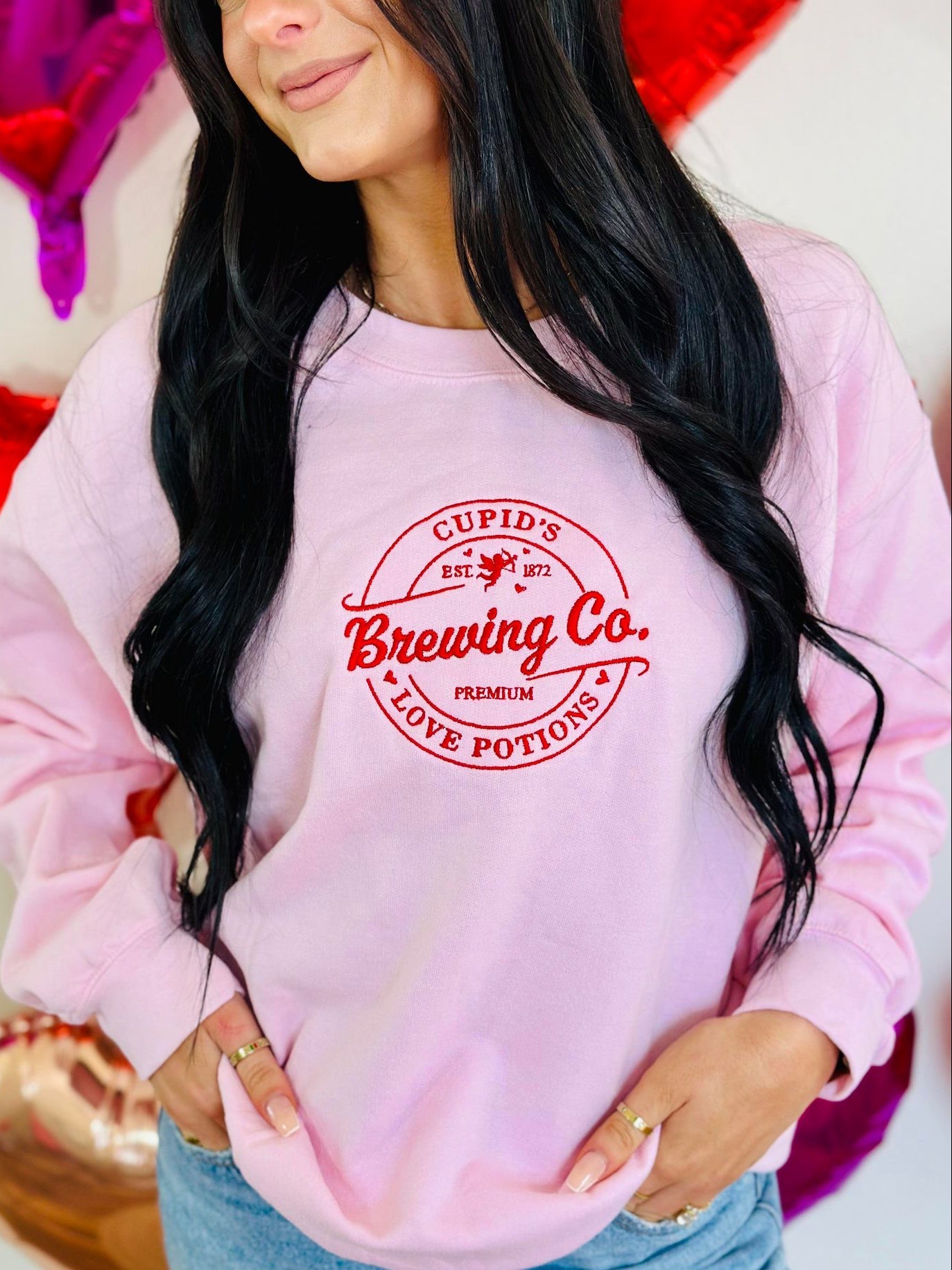 Person wearing a pink sweatshirt with 'Cupid's Brewing Co' print, surrounded by heart-shaped objects.