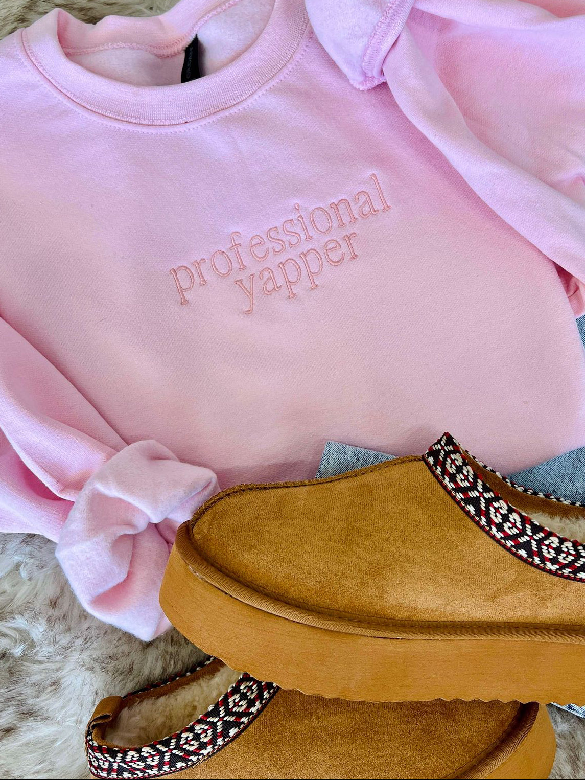 A light pink embroidered sweatshirt with the text 'PROFESSIONAL YAPPER' on the front, placed on a white furry surface, with a pair of blue jeans and a pair of tan casual shoes with embellished sole patterns on a matching white furry surface.