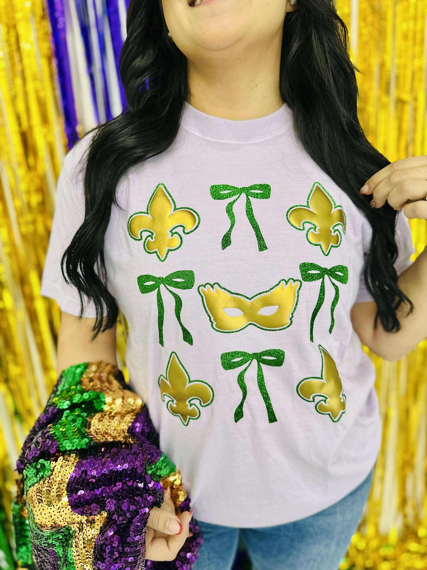 A person wearing a purple t-shirt with a green and gold bow design with masks and the text 'Mardi Gras' on it, standing in a glittery background.