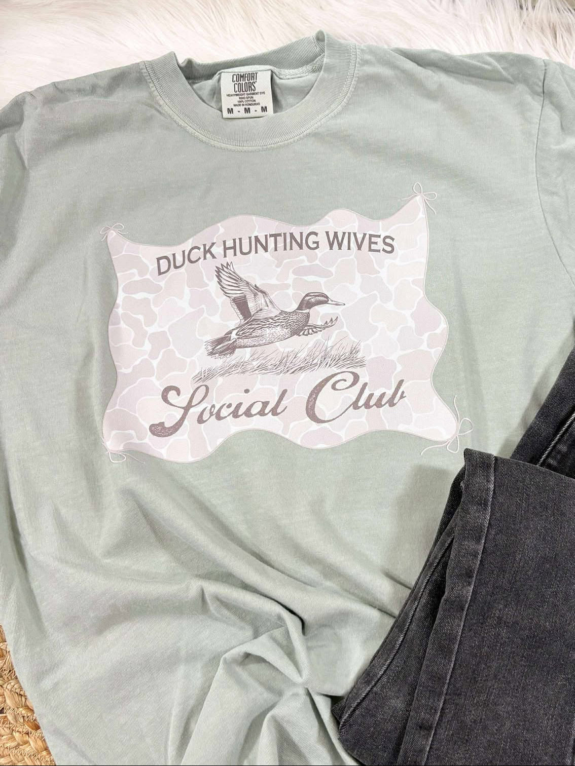 Light green t-shirt with Duck Hunting Wives Social Club graphic design on a textured surface