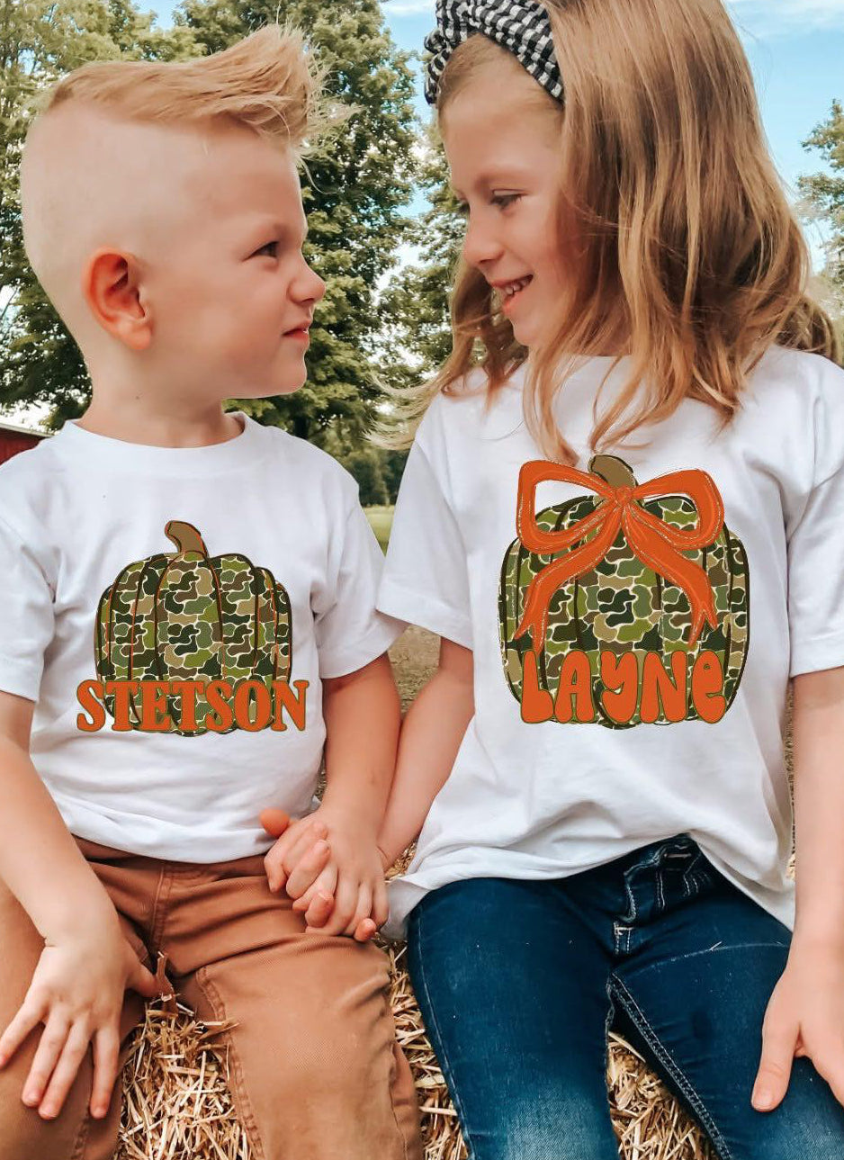 Two children wearing white t-shirts with custom pumpkin designs with names, sitting on a hay bale outdoors.