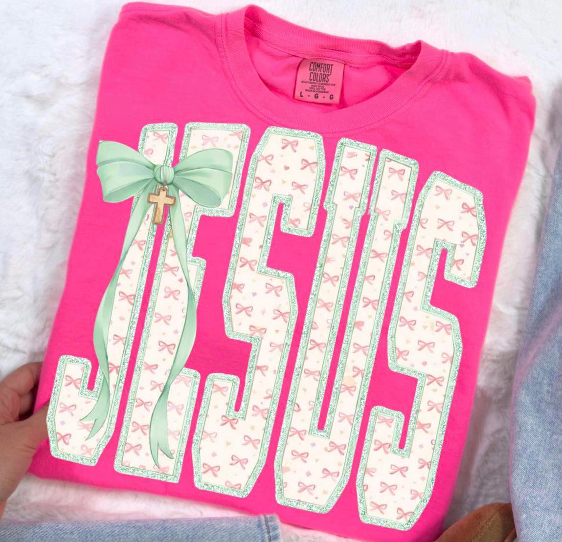 Pink t-shirt with 'Jesus' printed on it, featuring a green bow and cross charm, held by a hand.