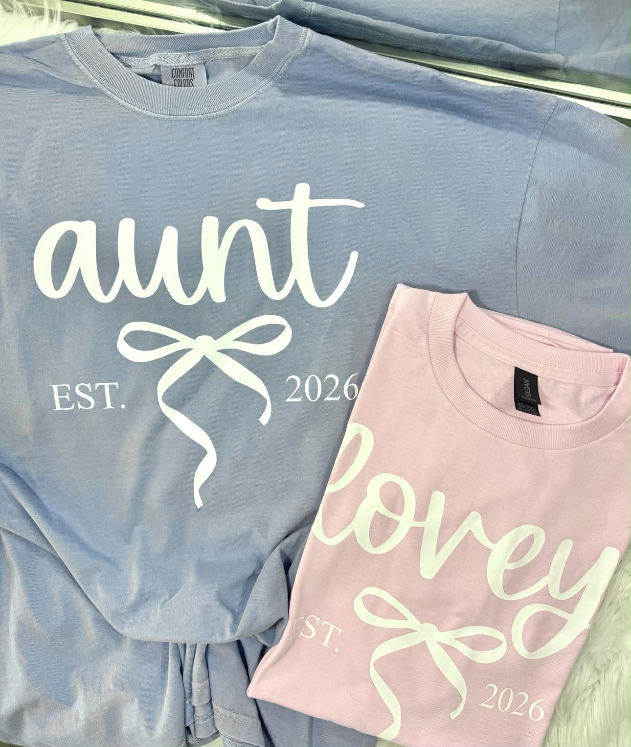 Two shirts, one blue and one pink, with 'aunt est. 2026' and 'lovey est. 2026' text.