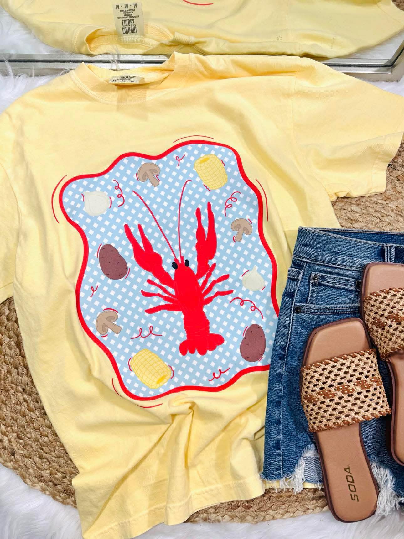 Yellow t-shirt with a red lobster design on a woven surface with jeans and sandals.