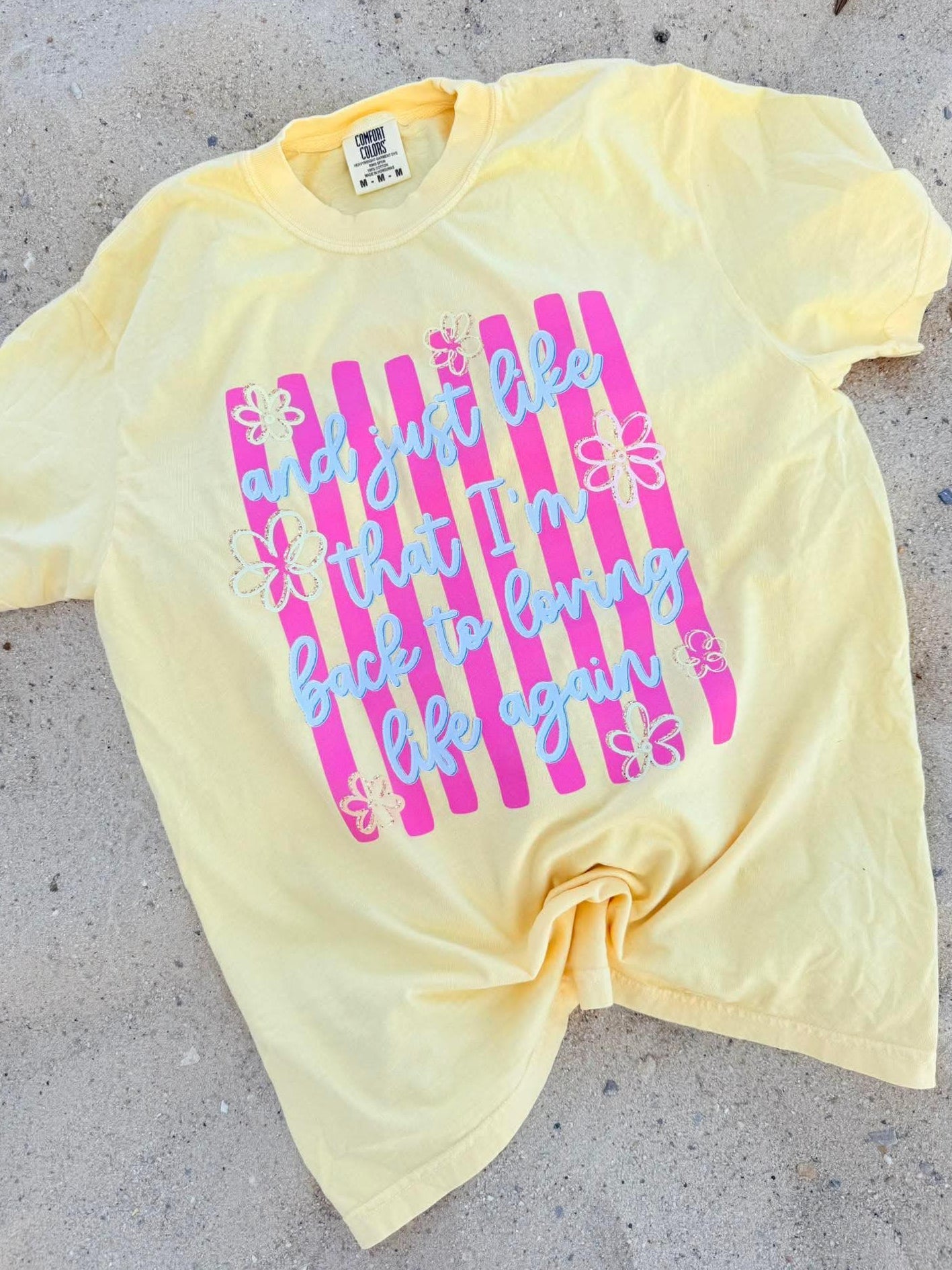 Yellow Butter t-shirt with pink and blue stripe "and just like that I'l back to loving life again" text on a beach surface