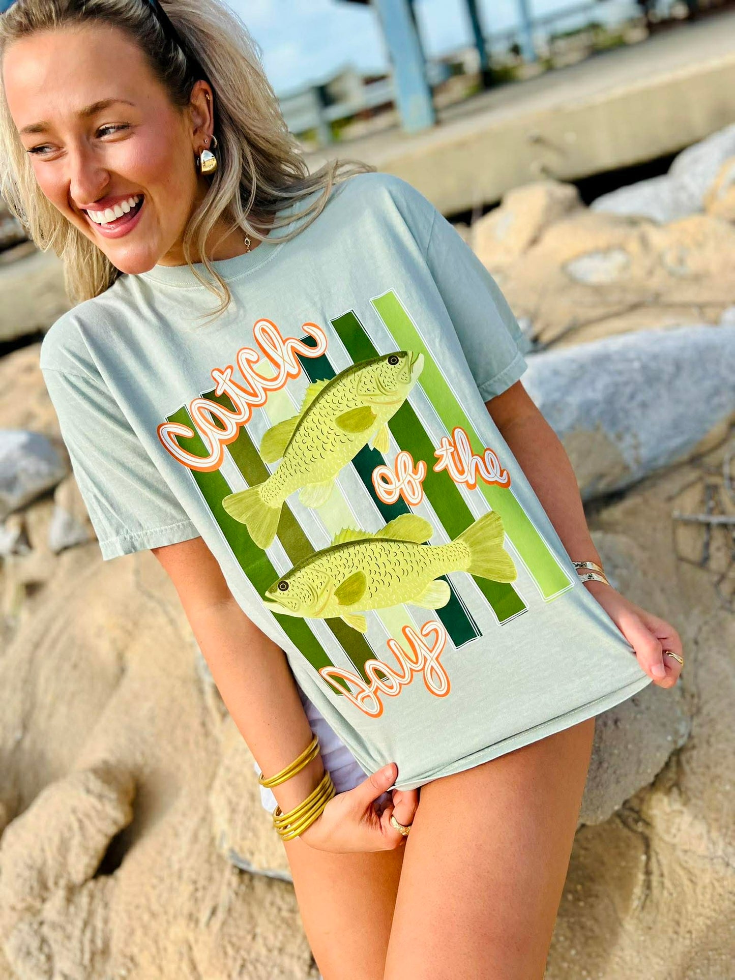 Woman wearing a light blue t-shirt with fish graphics and text on a rocky beach.