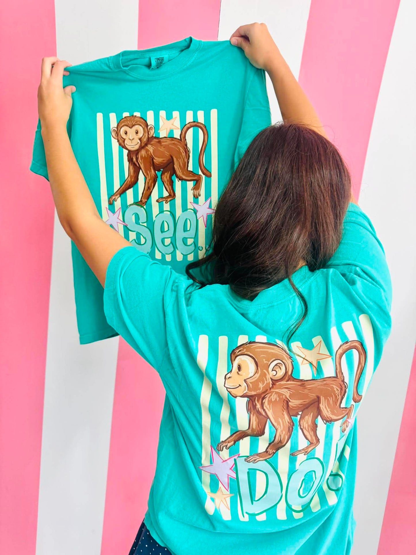 Person holding a turquoise t-shirt with monkey see monkey do striped graphics against a pink and white striped background