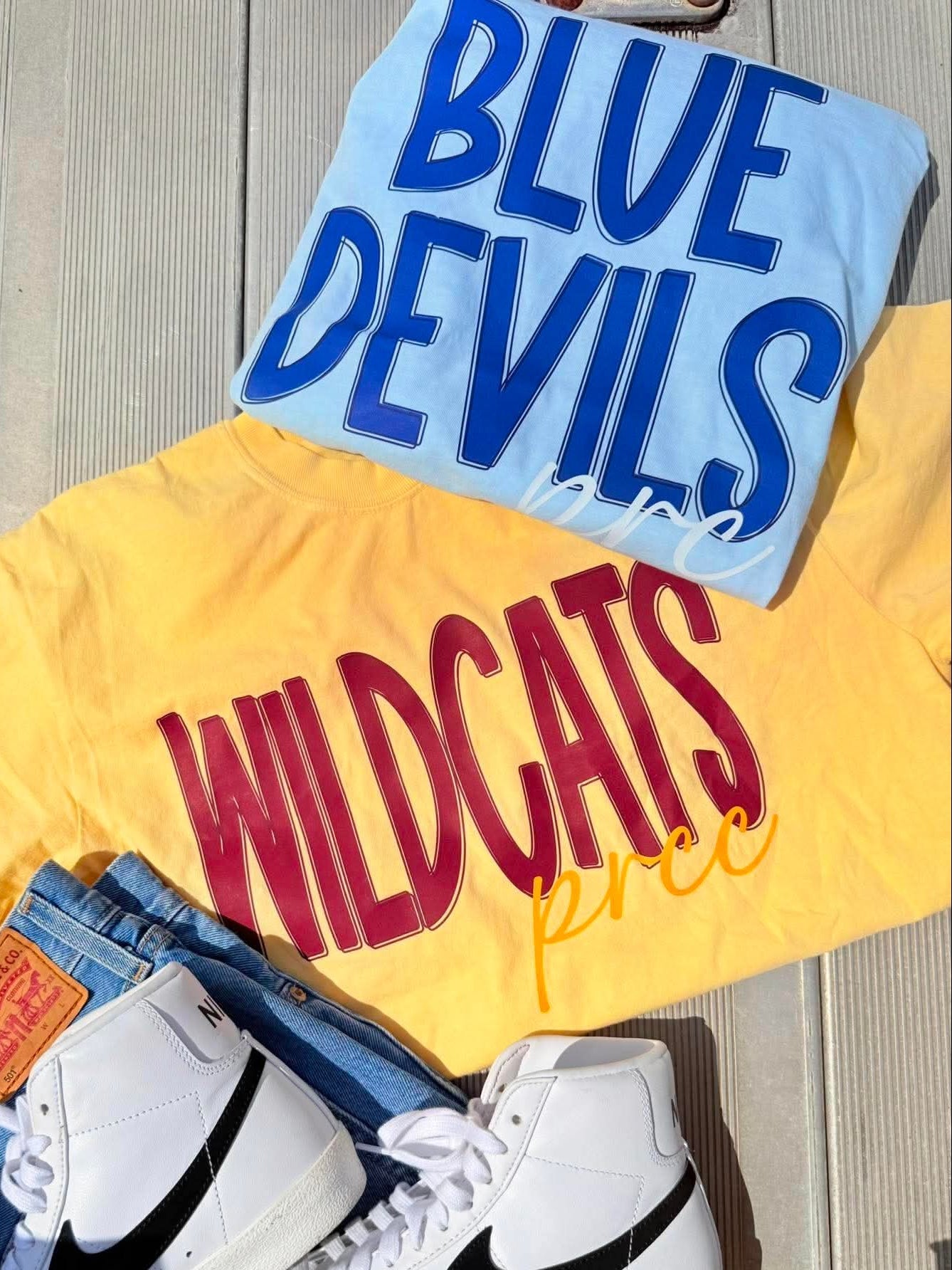 Yellow sweatshirt with 'WILDCATS' text, blue shirt with 'BLUE DEVILS' text, and white sneakers with black Nike logo on a wooden surface.