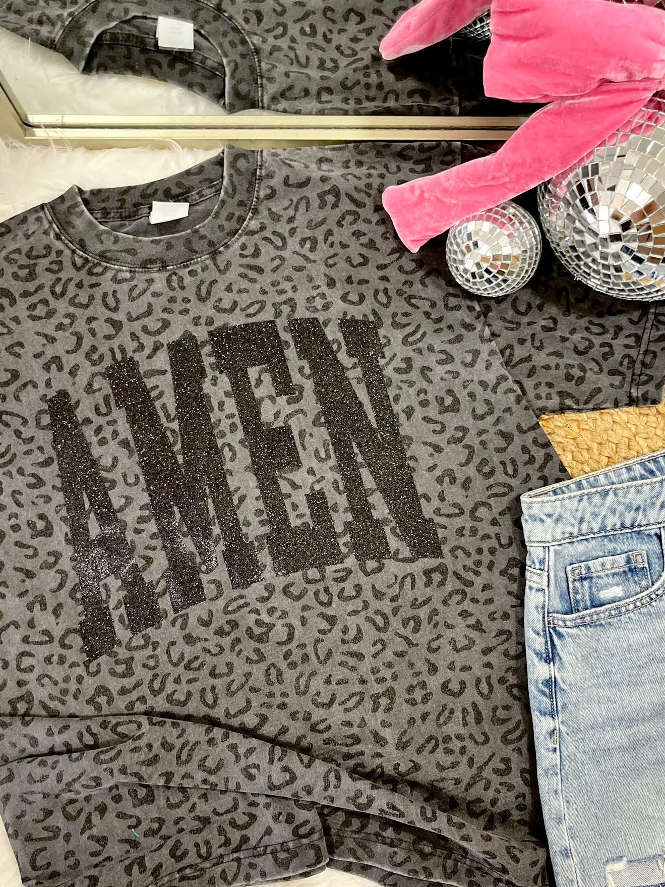 Gray leopard print sweatshirt with 'AMEN' glitter text, pink gloves, disco balls, and jeans on a surface.