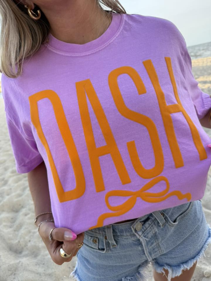 Person wearing a purple t-shirt with 'DASH' printed in orange on a beach.