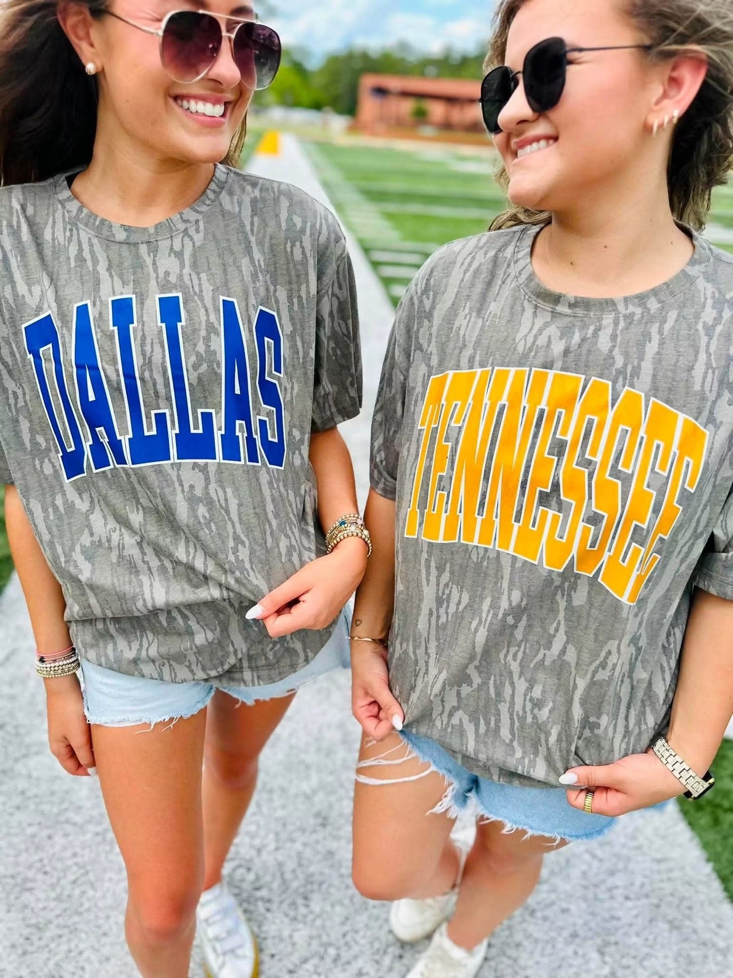 Two women walking outdoors wearing matching Mossy Oak Bottomland graphic t-shirts. The woman on the left has a blue 'DALLAS' print and the woman on the right has a yellow 'TENNESSEE' print, both paired with denim shorts and sunglasses.