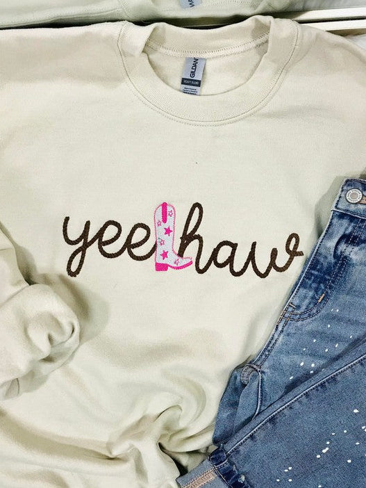 A tan embroidered sweatshirt with the text 'YEE HAW' on it, displayed on a surface with blue jeans and other clothing items.
