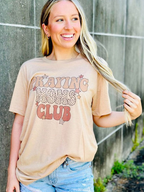 Woman wearing a beige t-shirt with text and denim shorts standing against a concrete wall Praying moms club.