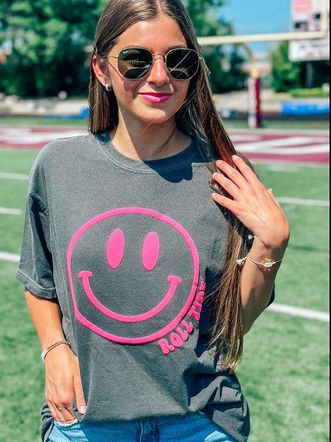 A female model wearing a gray t-shirt with a neon pink smiley face graphic on it, paired with denim shorts and mascot name, standing on an athletic field.