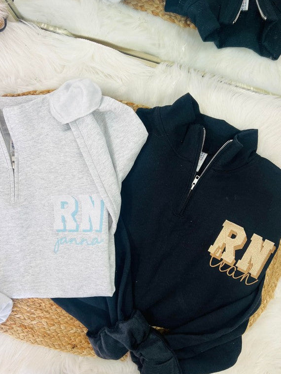 Two hooded jackets, one black and one gray, laid out on a white surface with 'RN' embroidered in gold on the front. Both jackets have a crew neck and a 1/4 zip design.