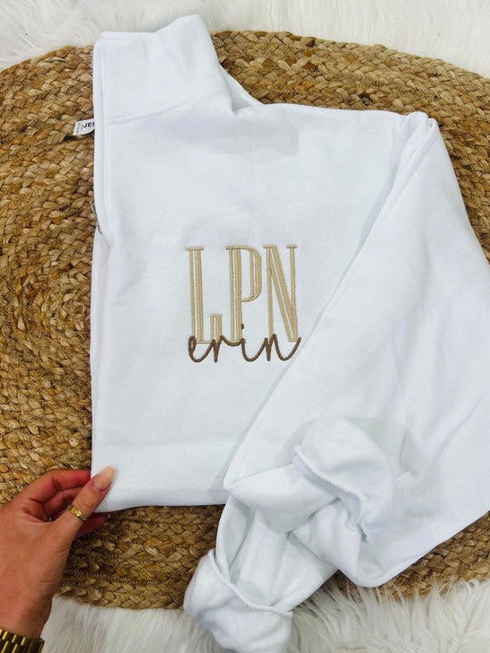 A white sweatshirt with the embroidered initial 'LPN' in brown on the chest area, displayed on a furry surface.