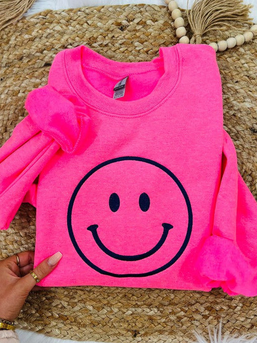 A neon pink sweatshirt with a black smiley face embroidered on the front, displayed on a furry surface.