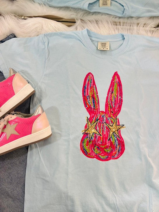 A light blue colored t-shirt with a graphic print of an eye-catching, colorful Easter bunny on it, displayed flat with a pair of pink sneakers and a white shoe on top of it.
