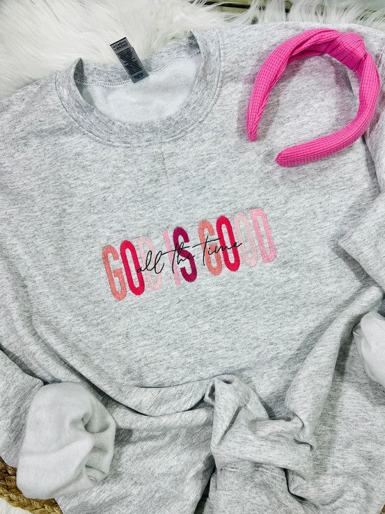 A gray sweatshirt with the phrase 'God Is Good' embroidered in pink on the front, displayed on a white fluffy surface with a pink hairband beside it.