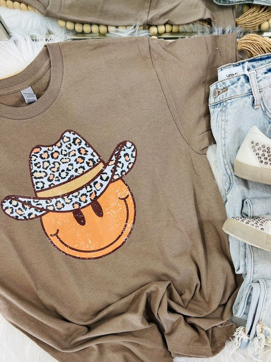 A brown short-sleeved graphic tee with a leopard wearing a cowboy hat and the text 'HAPPY' on it, displayed on a surface with jeans and shoes.