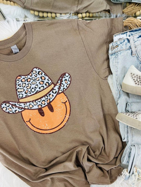 A brown short-sleeved graphic tee with a leopard wearing a cowboy hat and the text 'HAPPY' on it, displayed on a surface with jeans and shoes.