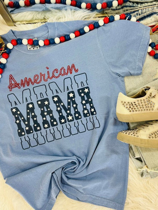 A blue t-shirt with the text 'American MAMA' printed on it, presented on a flat surface with decorative items around it.