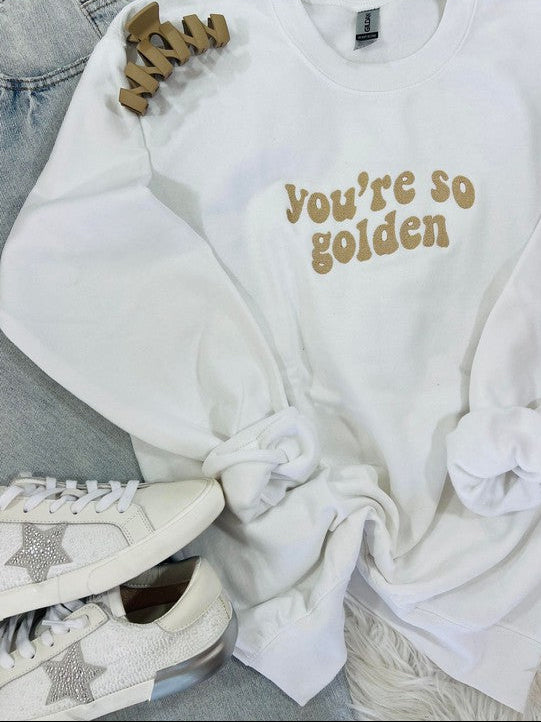 A white retro wave embroidered sweatshirt with the text 'you're so golden' on the front, displayed on a furry surface with a pair of sneakers and a star-shaped brooch.