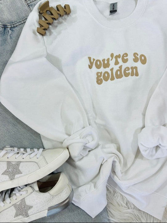 A white retro wave embroidered sweatshirt with the text 'you're so golden' on the front, displayed on a furry surface with a pair of sneakers and a star-shaped brooch.