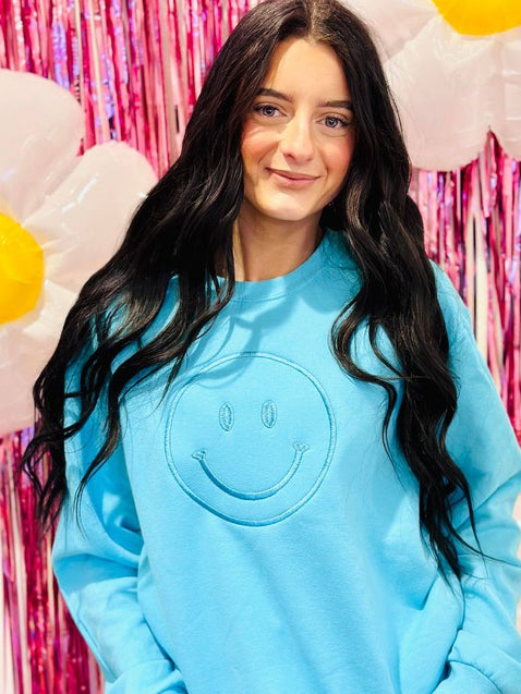 A person wearing a blue sweatshirt with a happy face embroidery on the front, posing against a pink glittery background with balloons.