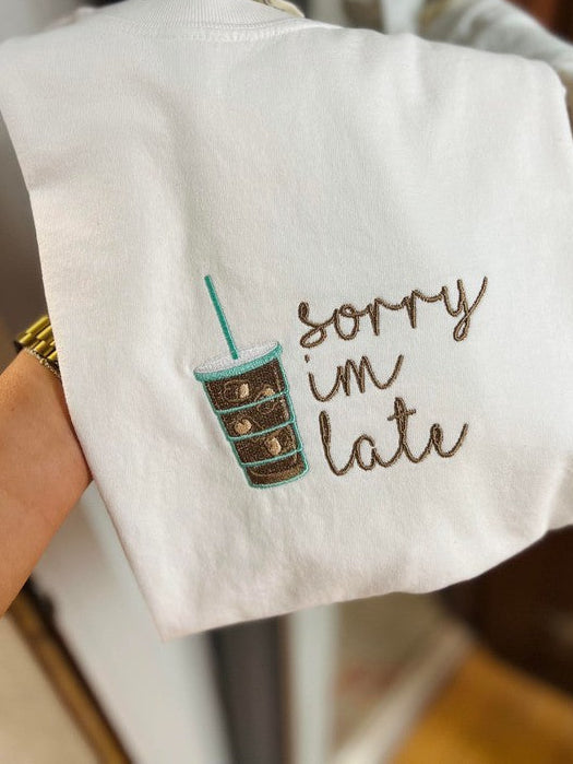 A white fabric with embroidered text 'Sorry I'm late' and a stylized iced coffee design held in a hand.