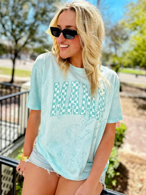 A woman wearing a light blue graphic tee with a checkered pattern, paired with shorts and sunglasses, standing near a metal railing.