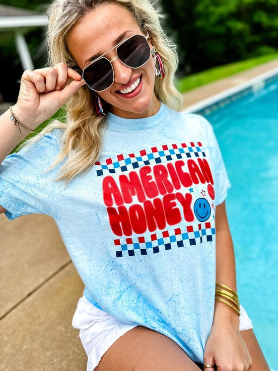 A person sitting by a poolside wearing a light blue graphic t-shirt with the text 'AMERICAN HONEY' in a patriotic design.