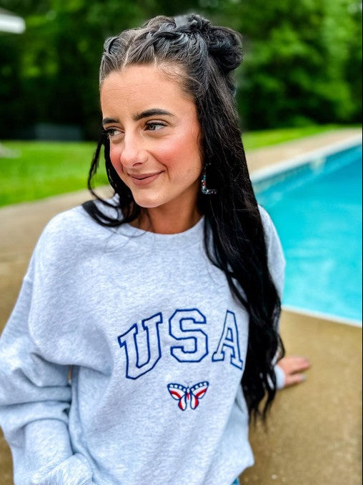 A person sitting by a poolside wearing an ash grey sweatshirt with the embroidered letters 'USA' and a butterfly logo on the front.
