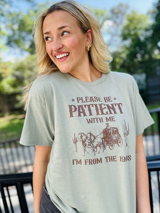 A woman wearing a green short-sleeved graphic tee with the text 'Please be patient with me I'm from the 1900's' printed on it, standing outdoors.