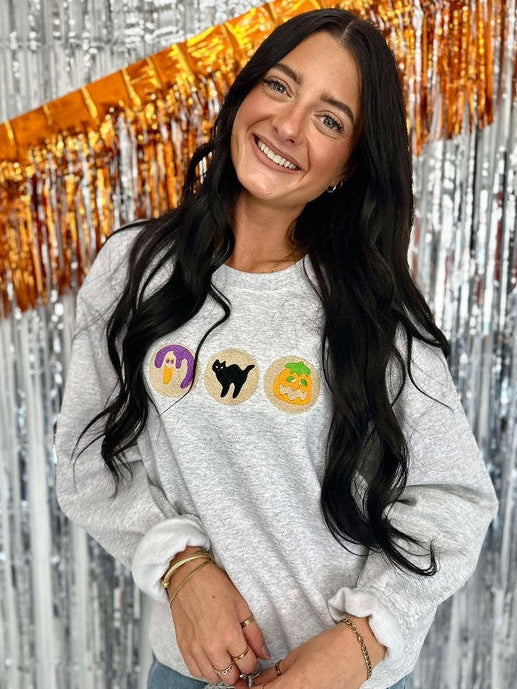 A woman smiling, wearing a long-sleeved gray sweatshirt with orange and black embroidered halloween cookies, and a sparkly background.