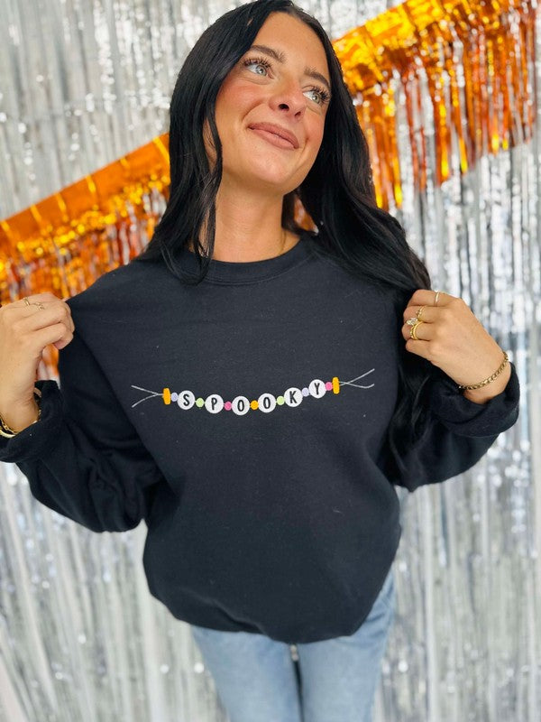A person wearing a black sweatshirt with 'Spooky Friendship Bracelet' embroidered on the front, posing against a glittery orange background.