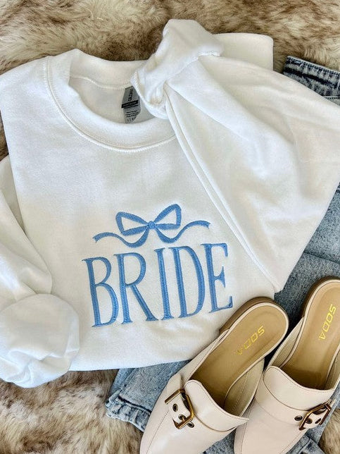 A white hoodie with the word 'Bride' embroidered on the front, paired with blue jeans and beige flats on a faux furry surface.