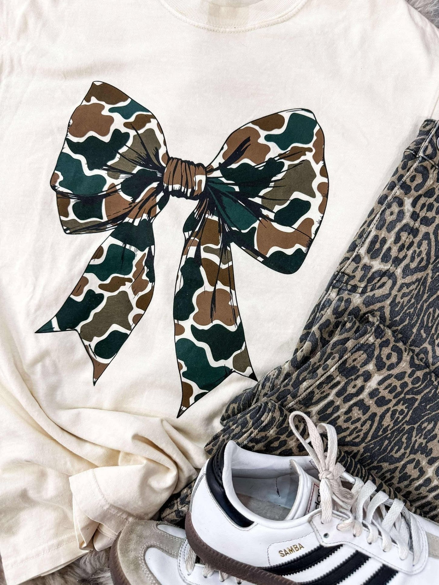 White sweatshirt with a large camouflage bow, leopard print pants, and Adidas sneakers on a white background.